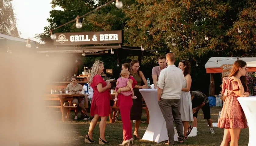 Ludzie rozmawiający przy stolikach i budce z napisem "Grill & Beer" na zewnątrz.